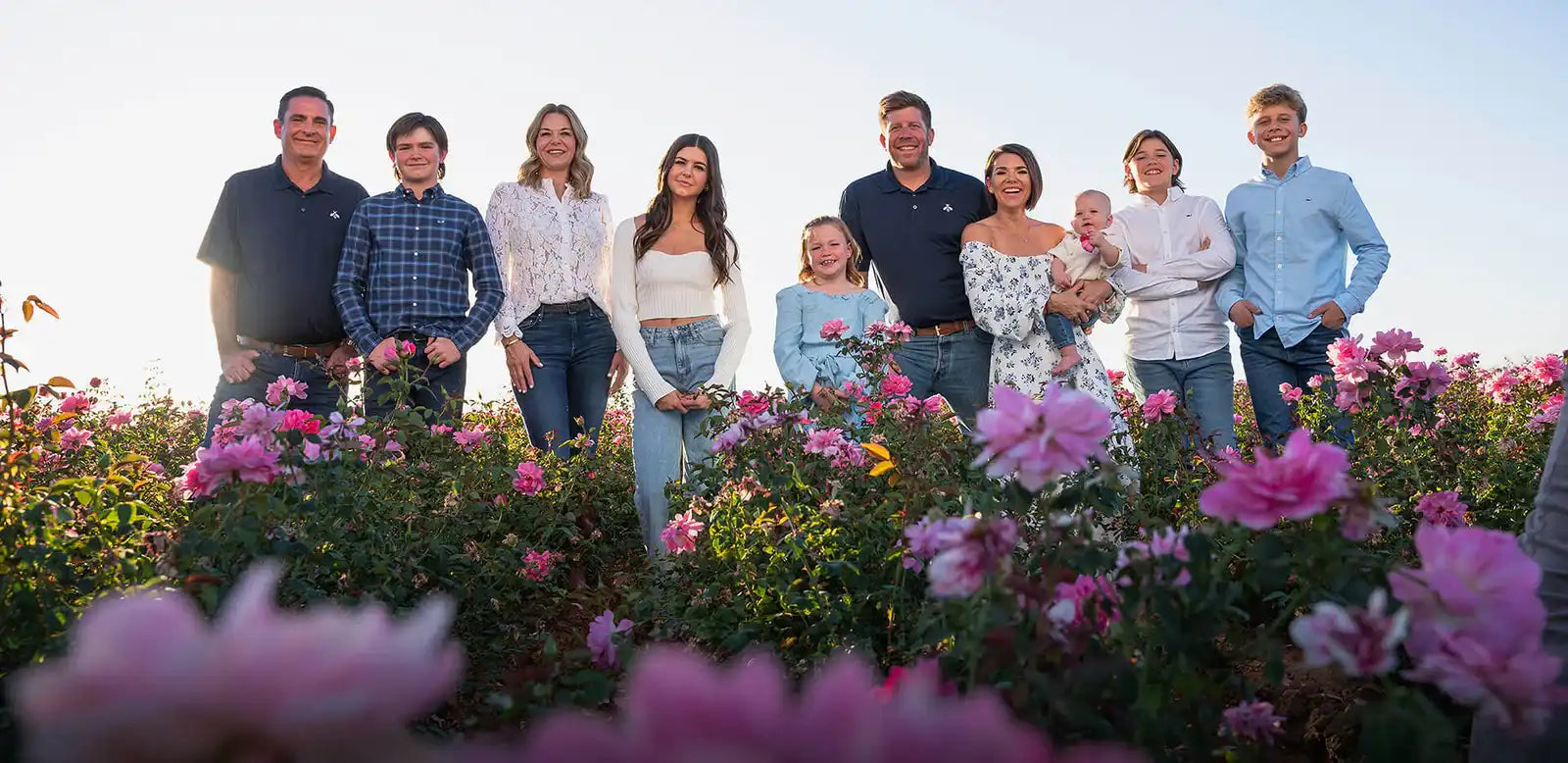 Family of nine standing in a field of pink flowers with a clear sky.