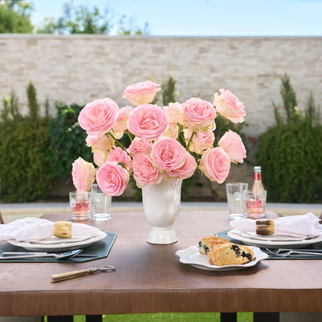 Bouquet of pink roses in a vase on a table with plates and glasses outdoors.