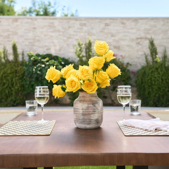 Yellow roses in a vase on a wooden table outdoors with glasses of water and white wine.