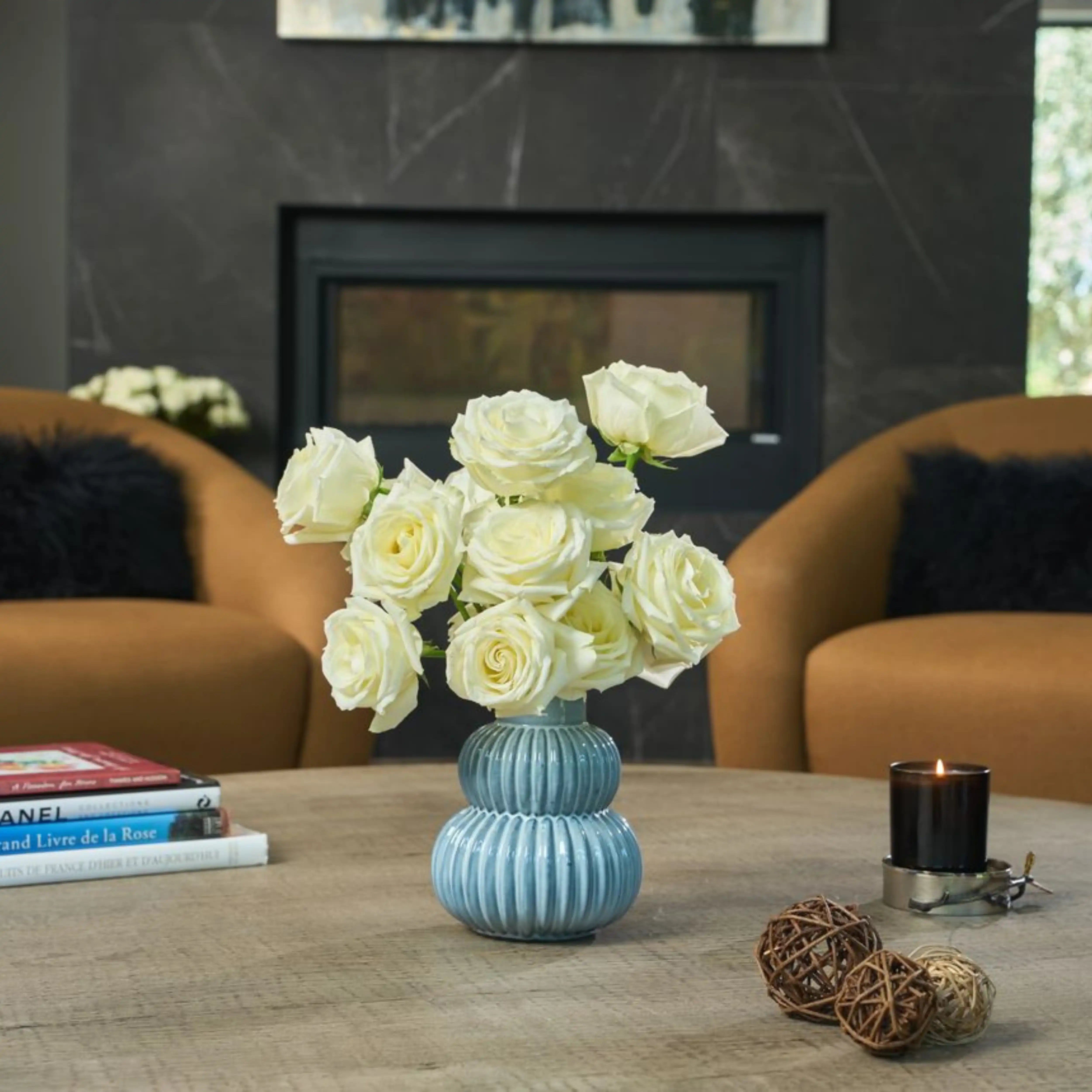 White roses in a blue vase on a coffee table with books and decorative items in a living room setting.