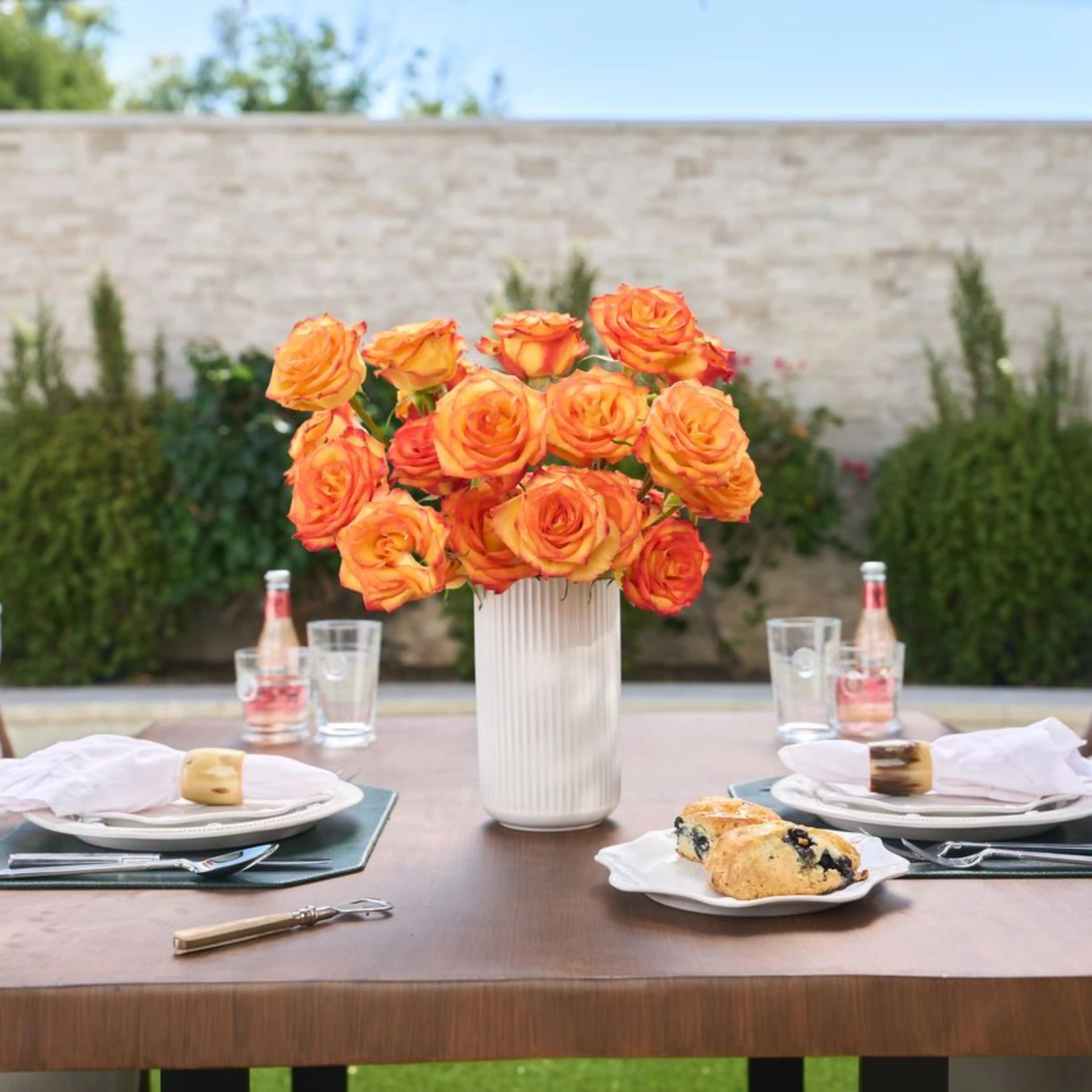 Outdoor table setting with orange roses in a vase, glasses, and pastries.