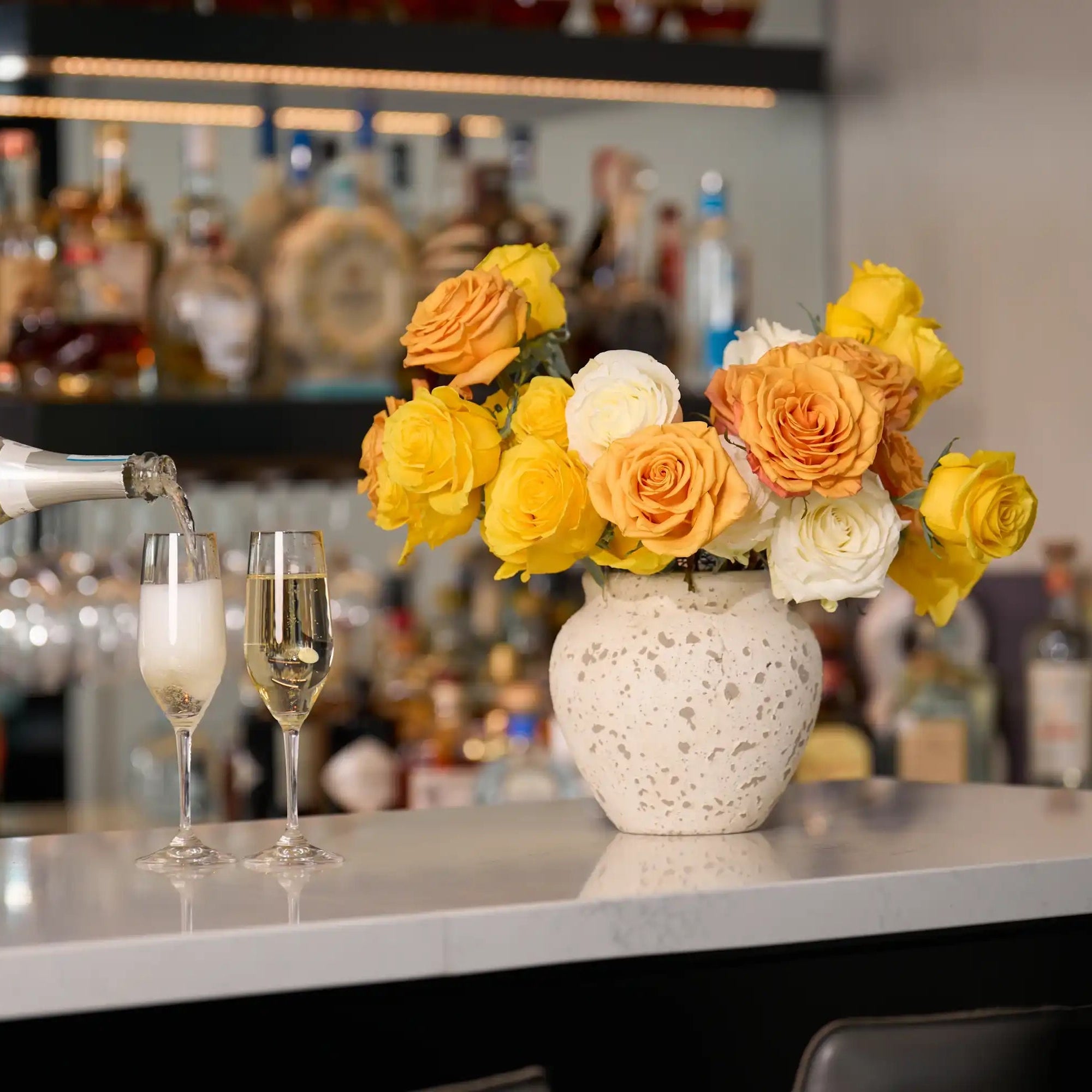 Person pouring champagne into a glass with a vase of flowers on a bar counter.