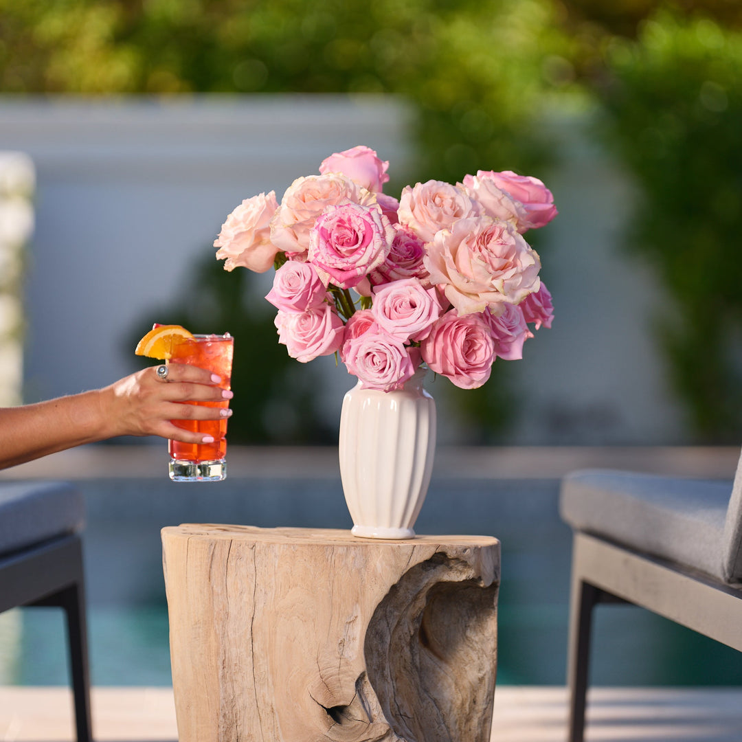 Person holding a drink with pink flowers on a wooden table outdoors.