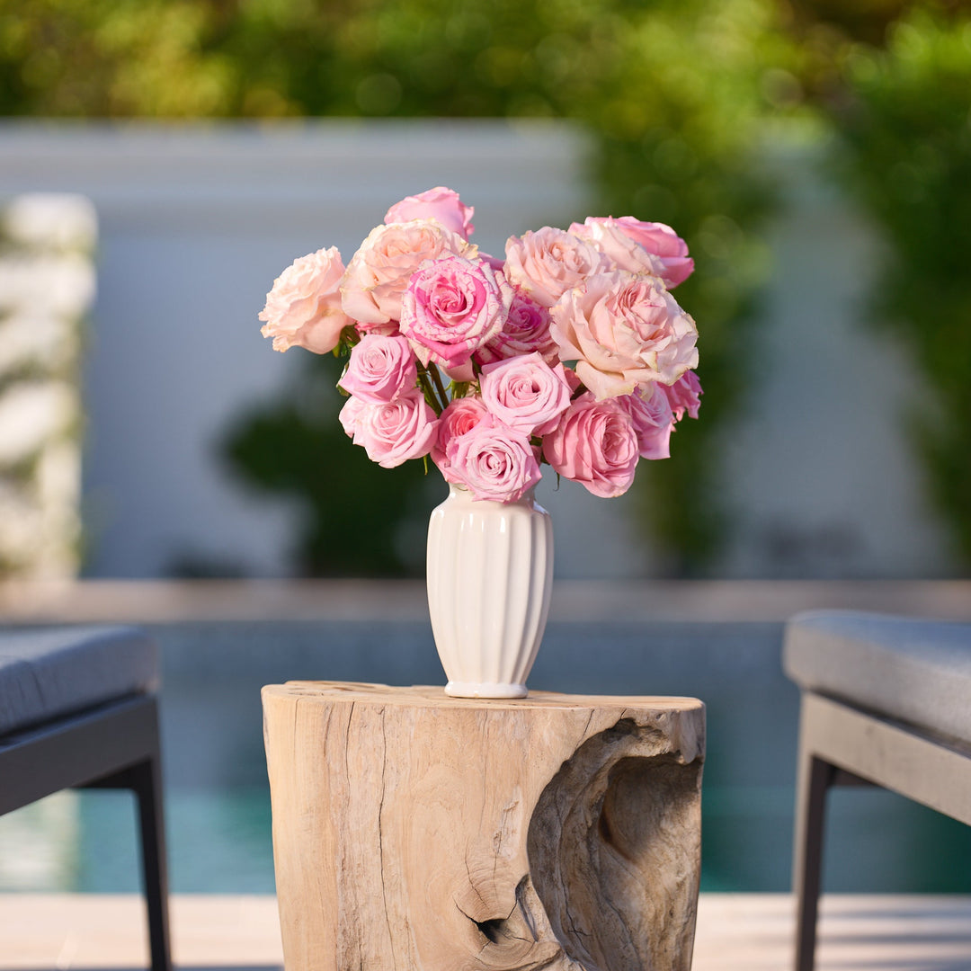 Bouquet of pink roses in a white vase on a wooden table with chairs and pool in the background