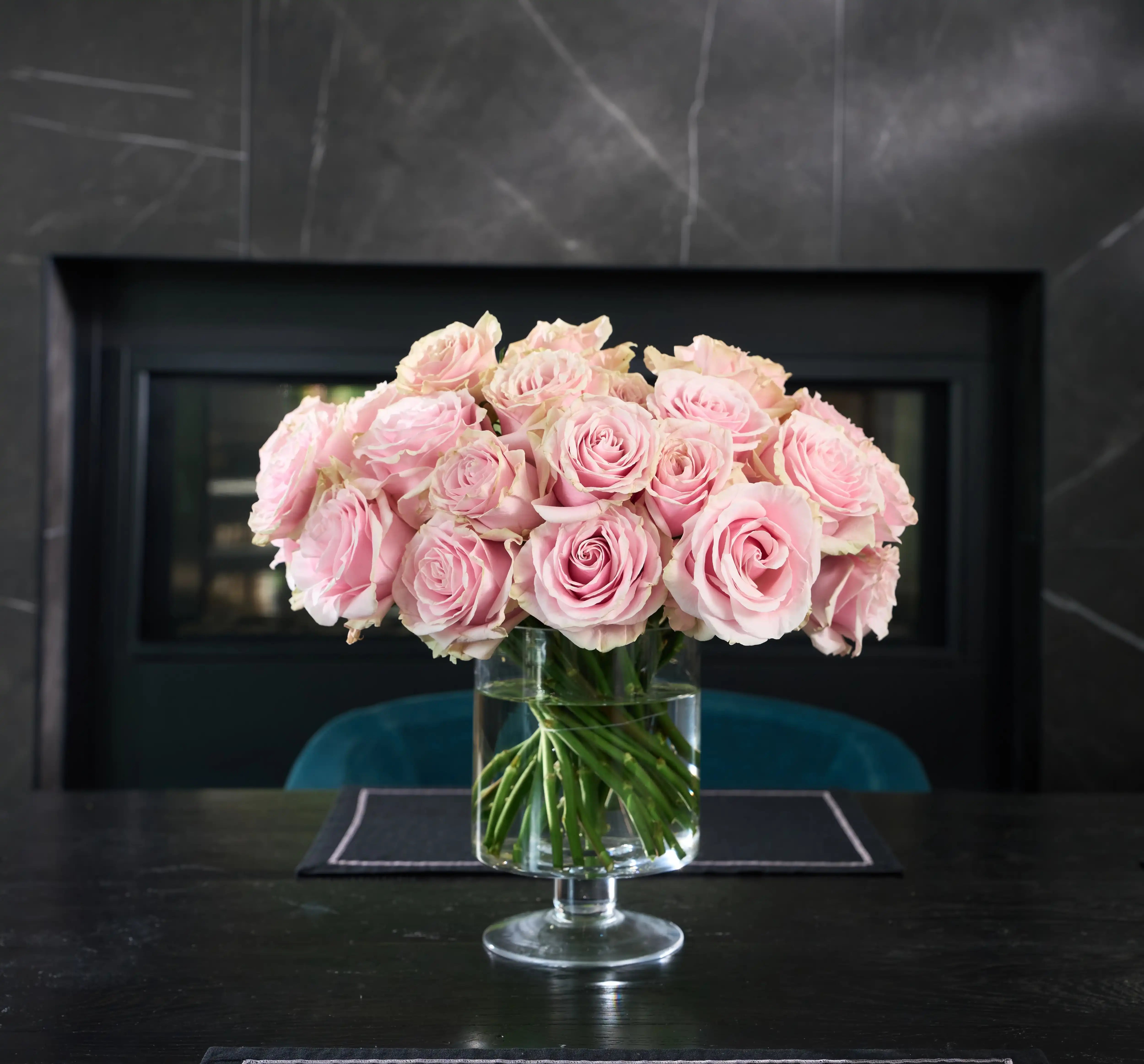 Bouquet of pink roses in a clear vase on a dark surface with a black fireplace in the background.