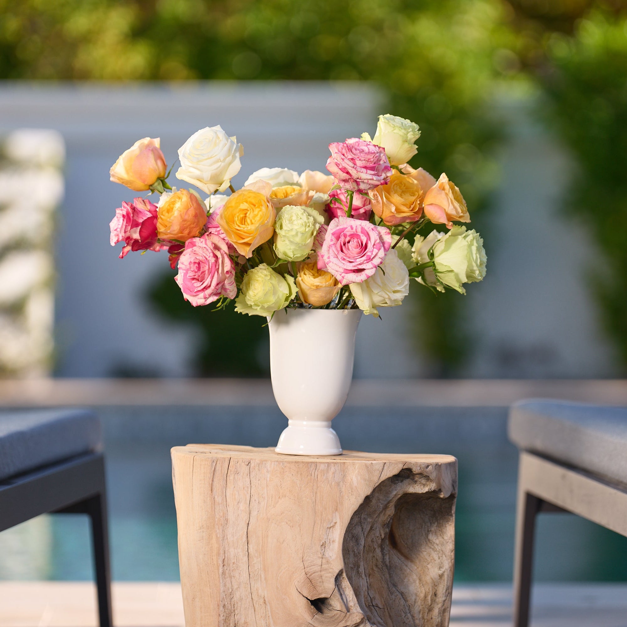 Floral arrangement in a white vase on a wooden table with chairs and blurred greenery in the background