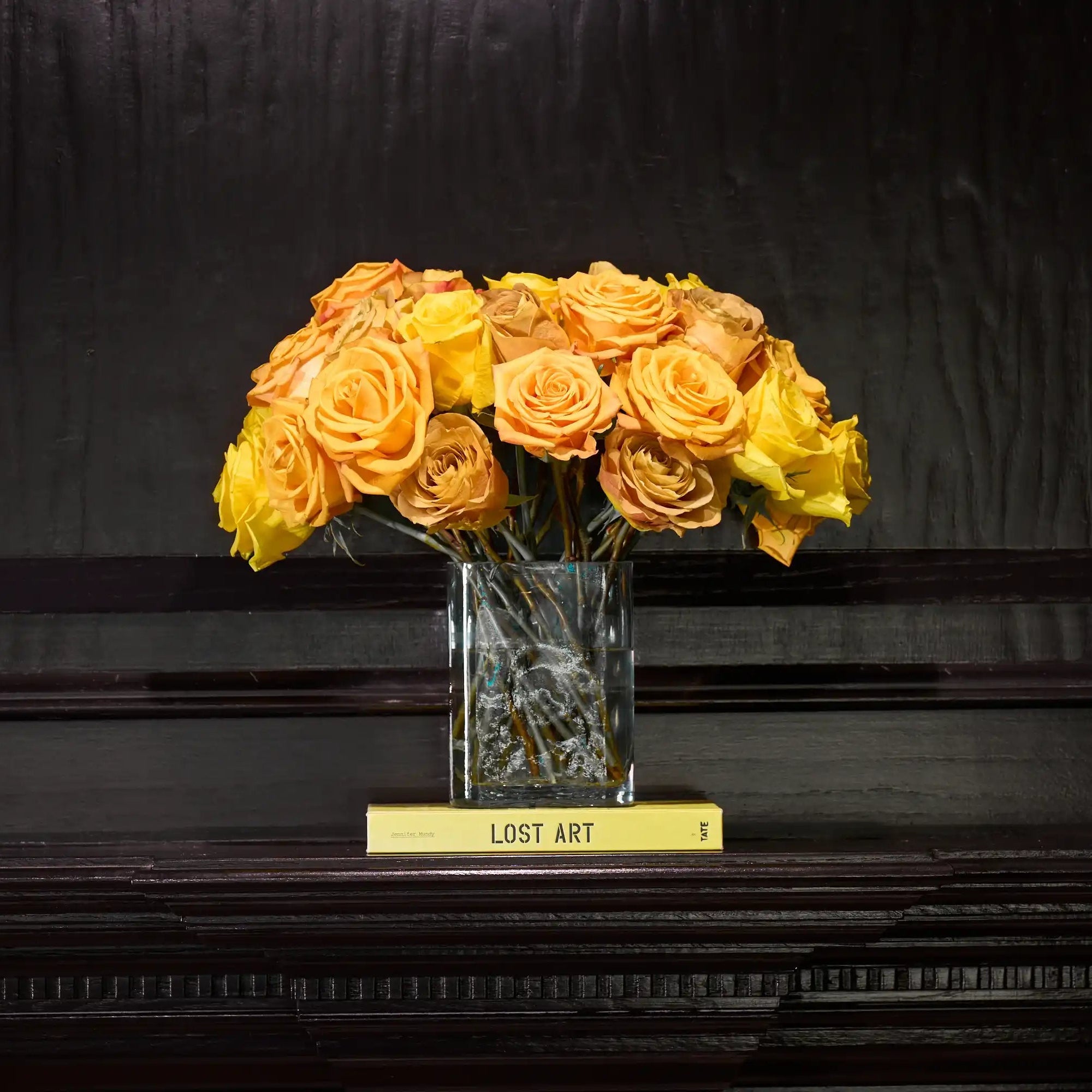 Bouquet of yellow roses in a clear vase on a black surface with 'Lost Art' book.
