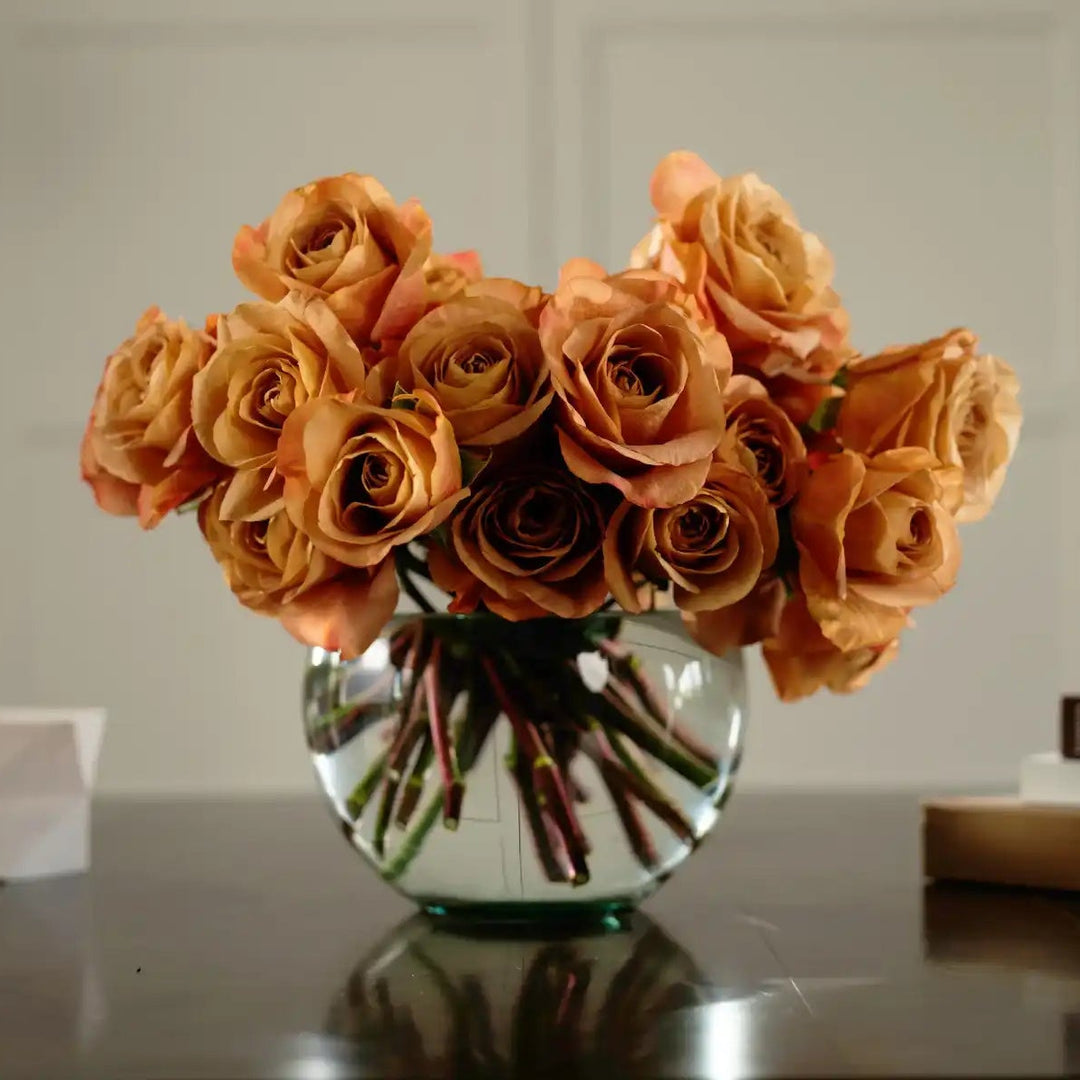 Bouquet of orange roses in a clear vase on a reflective surface with a neutral background